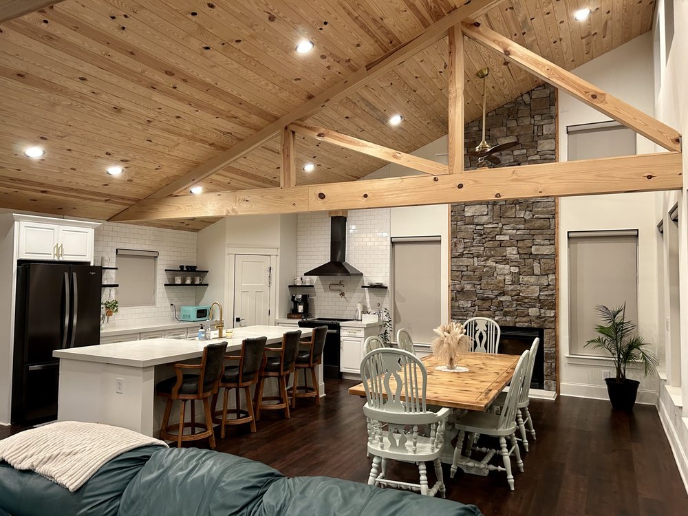 Open great room looking toward a white kitchen with vaulted beams and a stone fireplace