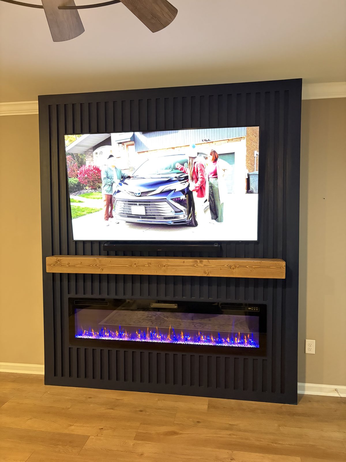 Dark vertical-slat fireplace and TV wall with a light wood mantel and linear electric insert