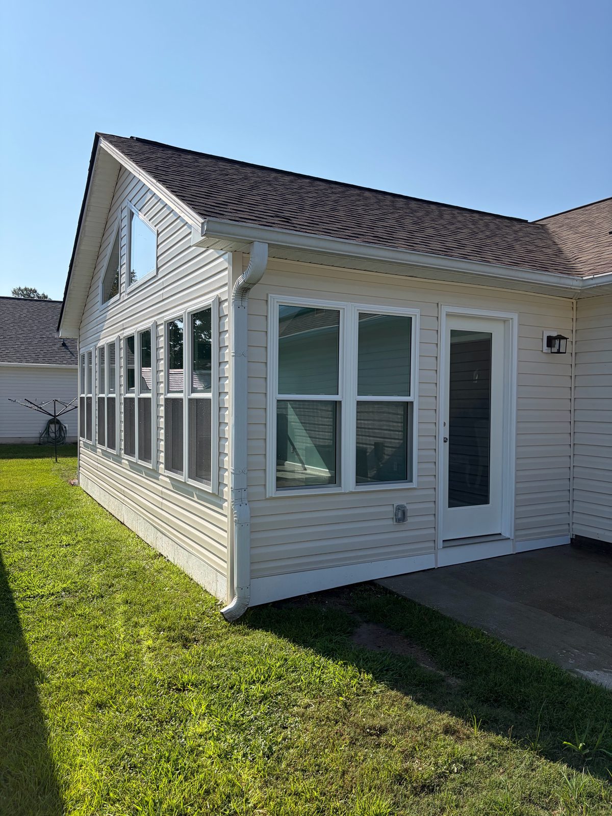 Gabled sunroom addition from outside with vinyl siding, a row of windows, and a door to the patio