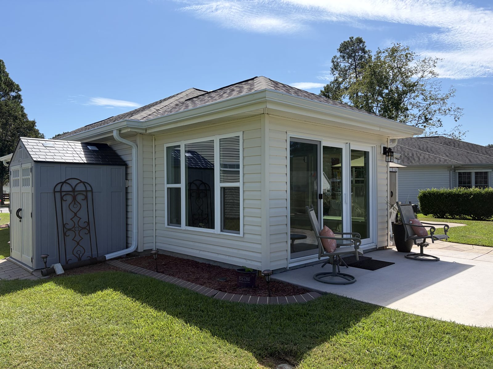 Hip-roof sunroom addition with vinyl siding, windows, and a sliding patio door opening onto a concrete patio, with a shed beside it