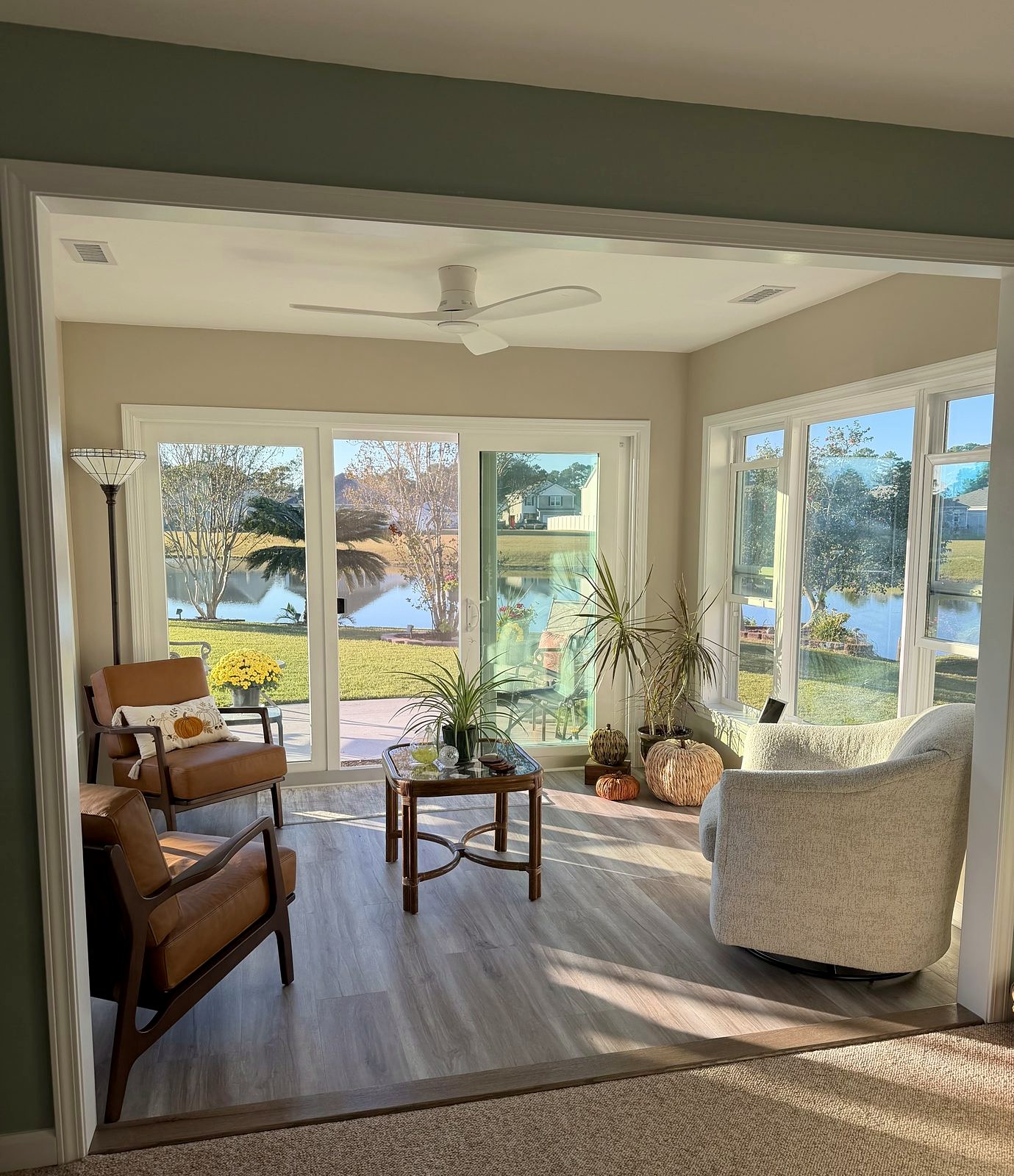Furnished sunroom addition in afternoon sun, with a sliding glass door and corner windows looking out on a pond
